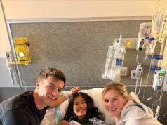 Young girl in hospital bed with two smiling visitors, medical equipment in background.