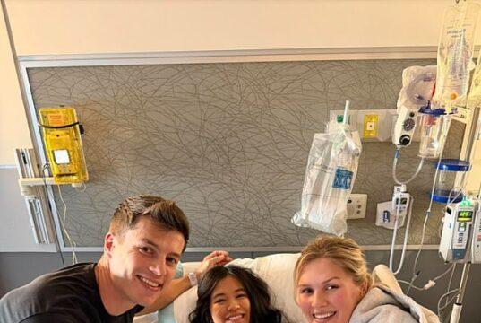 Young girl in hospital bed with two smiling visitors, medical equipment in background.