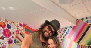 Huge man with curly hair, sunglasses, and a hat standing next to a smiling woman in black, vibrant colorful background.