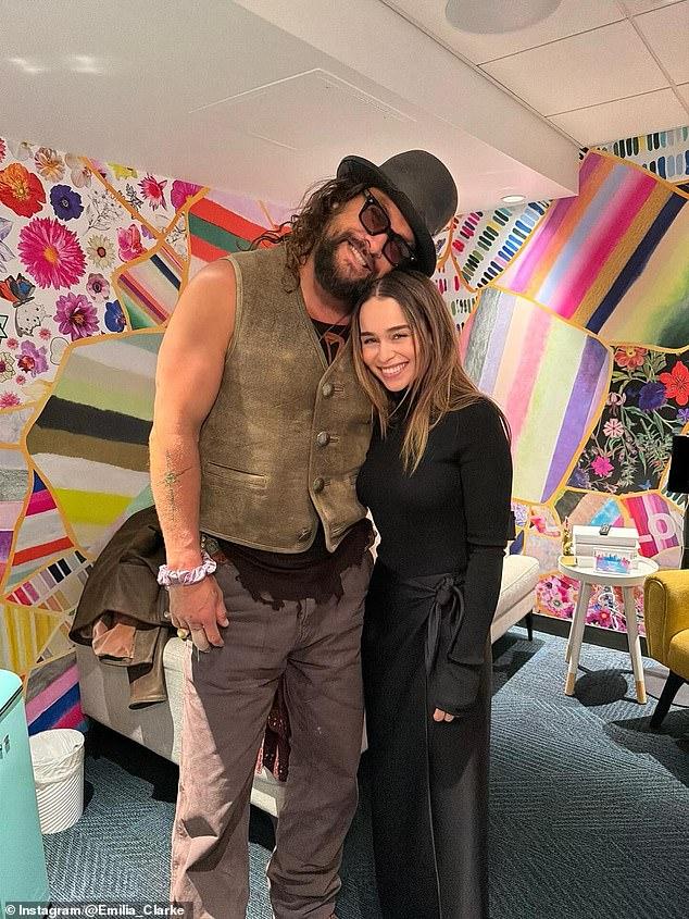 News Room : Game Of Thrones stars Jason Momoa and Huge man with curly hair, sunglasses, and a hat standing next to a smiling woman in black, vibrant colorful background.