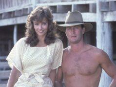 Young woman and shirtless man standing outdoors on wooden structure in vintage attire.