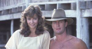 Young woman and shirtless man standing outdoors on wooden structure in vintage attire.
