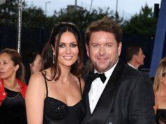 Elegant couple in formal black attire attending a glamorous event, surrounded by other guests.