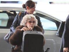 Woman with curly gray hair wearing sunglasses in a wheelchair at the airport, being assisted by a smiling staff member.