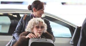 Woman with curly gray hair wearing sunglasses in a wheelchair at the airport, being assisted by a smiling staff member.