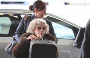 Woman with curly gray hair wearing sunglasses in a wheelchair at the airport, being assisted by a smiling staff member.