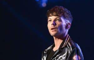News Room : Louis Tomlinson pays heartbreaking tribute to his One Direction bandmate Liam Payne in poignant song on his new album Young man performing on stage with microphone, dark background, casual stylish attire, tattoos on arm.