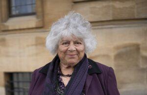 News Room : Miriam Margolyes lists stunning three-bedroom rural retreat in Australia for $4.2million Senior woman with curly gray hair smiling, standing outdoors in front of historic building, wearing purple jacket and scarf.