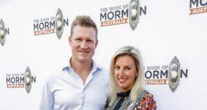 Elegant man and woman at book launch event for "The Book of Mormons Australia" in front of branded backdrop, red carpet, stylish attire, smiling, event photos.