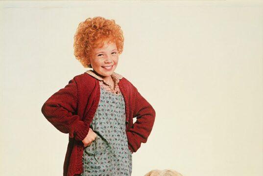 Adorable young girl with curly red hair in a vintage dress and cardigan standing next to a fluffy, friendly mixed breed dog.