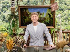 A young man in a light gray suit smiling at a desk decorated with wildlife sculptures outdoors.