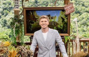 A young man in a light gray suit smiling at a desk decorated with wildlife sculptures outdoors.