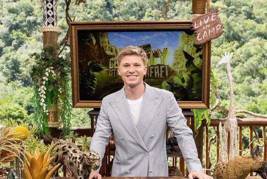A young man in a light gray suit smiling at a desk decorated with wildlife sculptures outdoors.
