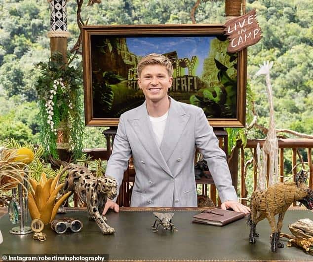 News Room : PETA's scathing letter to Robert Irwin ahead A young man in a light gray suit smiling at a desk decorated with wildlife sculptures outdoors.