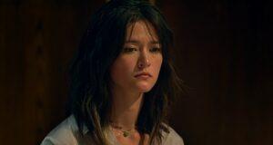 Young woman with dark wavy hair, contemplative expression, indoor dark background.