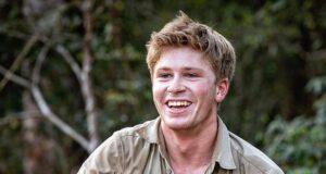 Young man smiling outdoors in khaki shirt at the zoo, holding a small item in his hand.