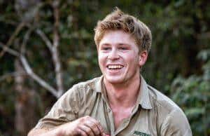 News Room : Robert Irwin caught off guard after radio host asks personal question Young man smiling outdoors in khaki shirt at the zoo, holding a small item in his hand.