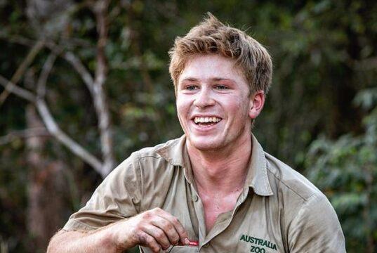 Young man smiling outdoors in khaki shirt at the zoo, holding a small item in his hand.
