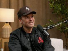 Young man with a microphone, wearing a New York Yankees cap, during podcast interview indoor setting with wooden decor and plants, smiling confidently.