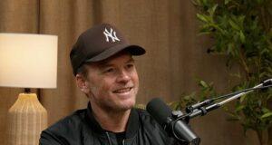 Young man with a microphone, wearing a New York Yankees cap, during podcast interview indoor setting with wooden decor and plants, smiling confidently.