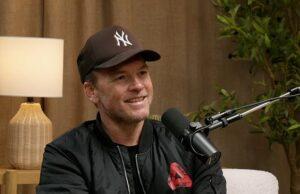 News Room : Sam Worthington says his bad attitude in an audition got him the lead role in Avatar: ‘This is a waste of time’ Young man with a microphone, wearing a New York Yankees cap, during podcast interview indoor setting with wooden decor and plants, smiling confidently.