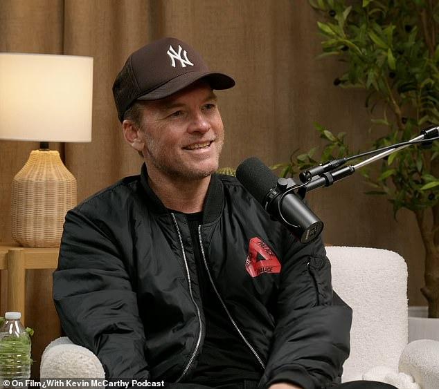 News Room : Sam Worthington says his bad attitude in Young man with a microphone, wearing a New York Yankees cap, during podcast interview indoor setting with wooden decor and plants, smiling confidently.