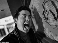 Young man making a funny face, sticking out his tongue near a large clock on a wall.