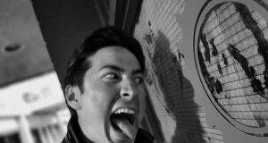 Young man making a funny face, sticking out his tongue near a large clock on a wall.