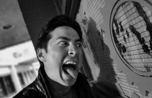 News Room : Secret woman in Heated Rivalry star Hudson Williams’ life… as unseen pictures from college days show heartthrob actor as you’ve never seen him Young man making a funny face, sticking out his tongue near a large clock on a wall.