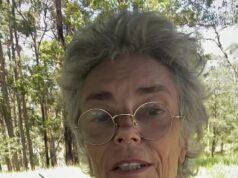 Elderly woman with glasses and floral blouse outdoors in sunny forest setting.