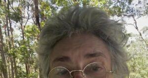 Elderly woman with glasses and floral blouse outdoors in sunny forest setting.