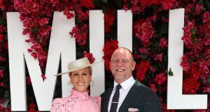 Elegant woman in pink dress with hat, standing with man in dark suit at floral backdrop event.
